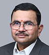 A middle-aged man with short black hair, glasses, and a mustache, wearing a dark suit, light shirt, and a small red mark on his forehead, poses against a plain gray background.